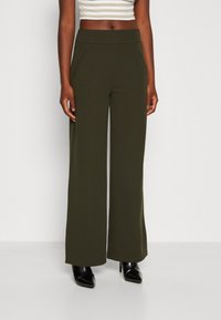 Olivengrüne weite Hosen aus dehnbarem Stoff mit hoher Taille und seitlichen Taschen, kombiniert mit schwarzen spitz zulaufenden Absätzen.