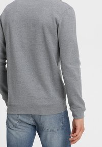 Sudadera gris hecha de tela suave, con mangas largas, cuello redondo y puños de canalé. Parcialmente metida dentro de unos jeans de mezclilla azules.