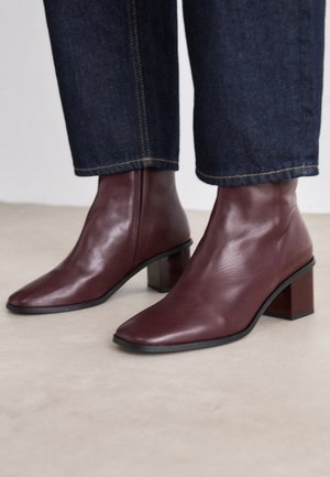 Knöchelhohe burgunderrote Lederstiefel mit Blockabsätzen, getragen zu dunkelblauen hochgekrempelten Jeans auf hellem Boden.