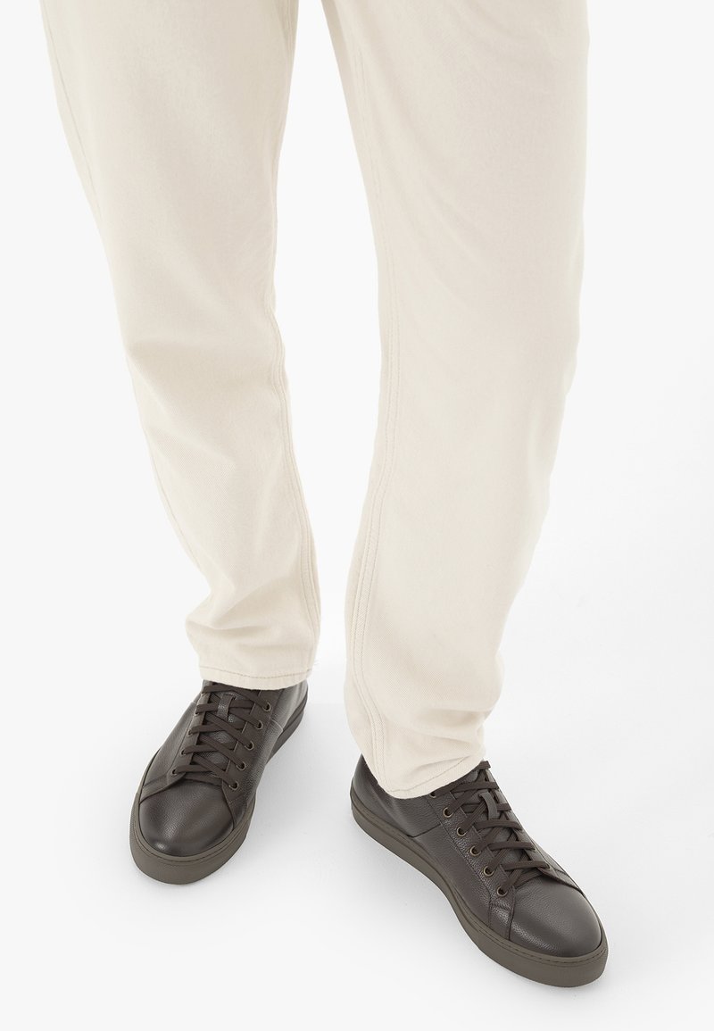 Personne portant un pantalon écru et des baskets en cuir marron foncé à lacets, debout sur un fond blanc.