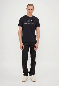 Vīriešu models, kurš valkā melnu Armani Exchange T-kreklu, melnas bikses un baltas sneakers, stāv pretī vienkāršai gaišai fona.