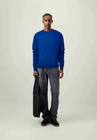 Homem a usar um suéter azul brilhante e jeans cinzentos desbotados, segurando uma mala preta, de pé contra um fundo claro e liso.