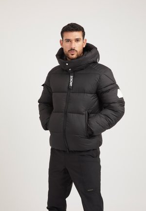 Homme portant une doudoune noire à col montant et un pantalon noir assorti, debout, les mains dans les poches, sur un fond uni.