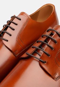 Scarpe eleganti in pelle marrone con finitura lucida, dotate di lacci marroni scuri e cuciture a contrasto. Texture liscia, design con punta rotonda.