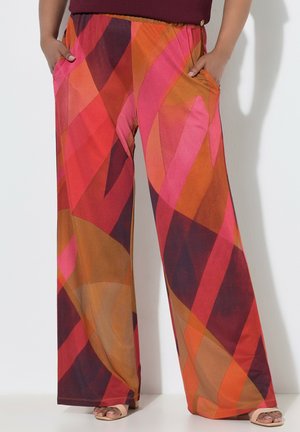 Pantalon à jambes larges avec des motifs géométriques audacieux dans des tons de rouge, orange, rose et marron, porté avec les mains dans les poches et des chaussures ouvertes beige.