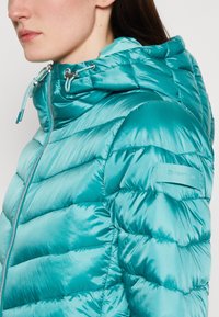 Doudoune turquoise avec capuche, présentant une finition brillante, un design matelassé et des cordons de serrage ajustables avec des embouts en métal.