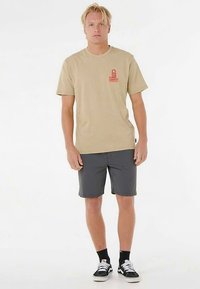 Beiges T-Shirt mit kurzen Ärmeln und einem roten Logo auf der Brust, kombiniert mit dunkelgrauen Shorts und schwarzen Turnschuhen. Einfacher, lässiger Schnitt.