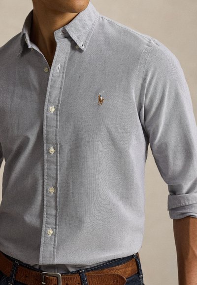 Man wearing light gray button-up shirt with rolled sleeves, brown belt, and embroidered polo player logo on left chest.