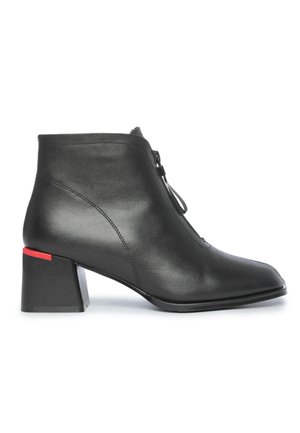 Bottines en cuir noir avec fermeture éclair frontale et talons blocs portées sur des jambes nues sur un fond clair.