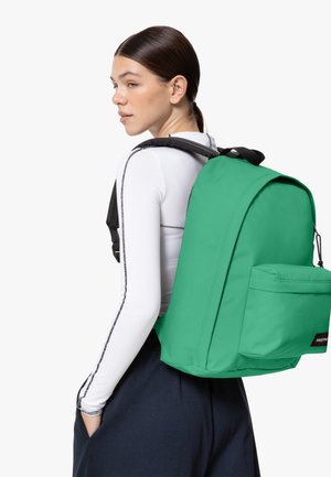 Green backpack with a structured shape, made of durable fabric. Features a front pocket and adjustable straps, seen on a person wearing a white top.