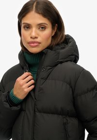 Zwarte pufferjack met een hoge kraag en afneembare capuchon. Voorzien van een ritsluiting en elastische manchetten. Het model draagt een groene gebreide sweater eronder.