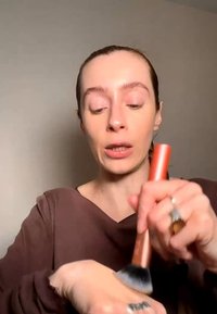 Femme aux cheveux tirés en arrière fait tourner un pinceau de maquillage à manche orange sur le dos de sa main, portant un haut marron sur un fond uni.