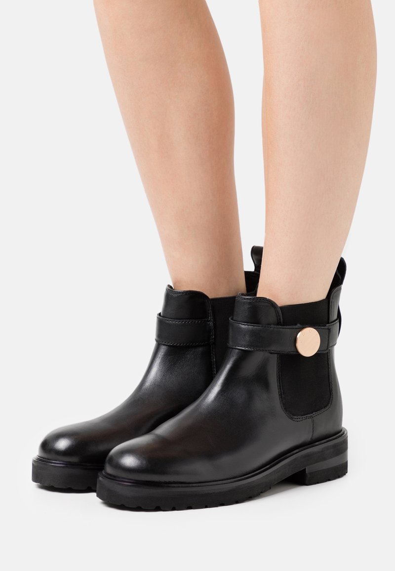 Bottes chevilles en cuir noir à finition lisse, avec des panneaux latéraux élastiques, un accent métallique rond et une semelle en caoutchouc texturée et basse.