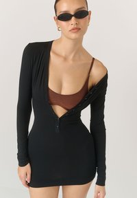 Czarna mini sukienka z długim rękawem i głębokim dekoltem w kształcie litery V, z detalami w postaci guzików oraz dopasowanym krojem; nałożona na brązowy biustonosz.