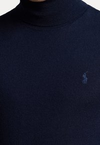 Laivastonsininen neulottu pooloneule pehmeästä kankaasta, jossa on resorireunainen kaulus ja hillitty kirjailtu logo rinnassa.