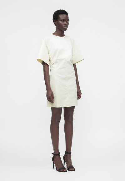 Γυναίκα όρθια, φοράει κρεμ φόρεμα με κοντά μανίκια μέχρι το γόνατο με πτυχώσεις στα πλευρά και μαύρα ψηλοτάκουνα σανδάλια, με φόντο απλό.