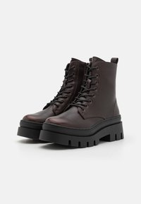 Botine din piele maro până la gleznă, cu talpa neagră groasă, șireturi și manșetă de prindere. Textură netedă, cu detalii cosute pe margini.