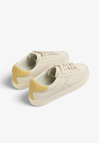 Sneakers in suede beige con un accento giallo sul tallone, dotati di lacci piatti, suole in gomma testurizzata e un logo impresso sul lato.