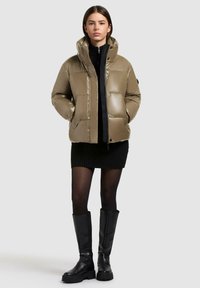 Giacca kaki imbottita con colletto alto, zip nera a contrasto e texture lucida. Indossata sopra un vestito nero a coste con collant texturizzati.