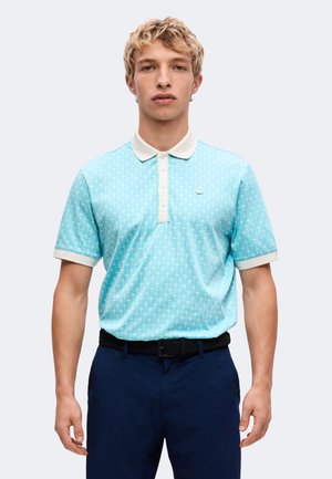 Ung mand iført en lyseblå mønstret poloshirt med hvid krave og mørkeblå bukser, stående mod en ensfarvet lys baggrund.