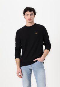 Zwarte gebreide trui met lange mouwen, ronde hals en een klein merklabel op de borst. Gecombineerd met lichtblauwe jeans.