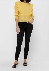 Gelbe Bluse mit gepunktetem Muster und Rüschen an den Schultern, kombiniert mit schwarzen Skinny-Jeans und schwarzen Riemchensandalen. Glatter Stoff und taillierte Passform.