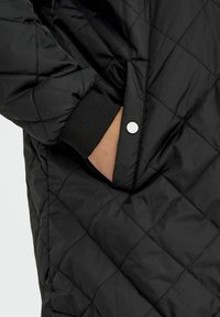 Schwarze gestepfte Jacke mit Rautenmuster, elastischen Bündchen und einer seitlichen Tasche mit Druckknopfverschluss. Glatte, glänzende Stofftextur.