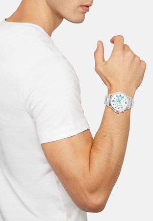 Sølv rustfrit stål ur med en hvid og turkis urskive, store tal og et struktureret rem, båret på et håndled iført en tætsiddende hvid skjorte.