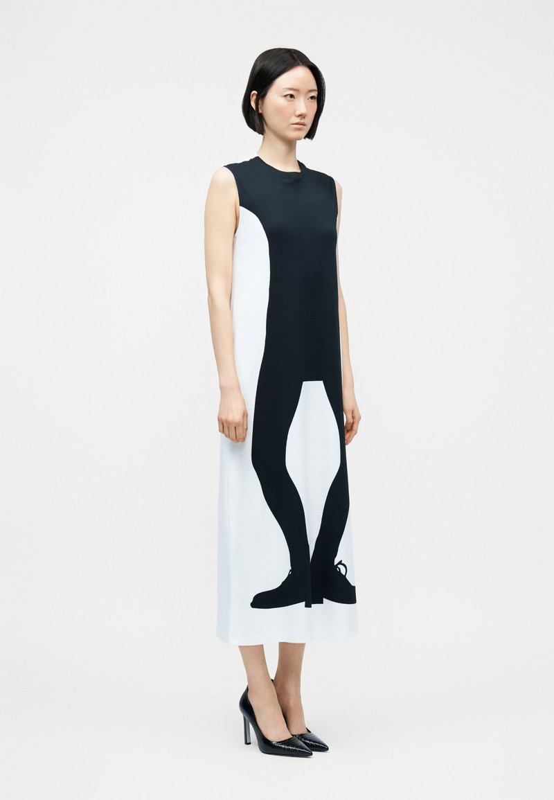 Femme portant une robe midi sans manches noire et blanche avec une impression graphique de jambes et de chaussures noires sur le devant, associée à des talons noirs.