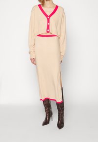 Beige cropped cardigan yang memiliki trim pink, dilengkapi dengan tiga kancing, dipadukan dengan rok midi yang serasi. Model mengenakan sepatu bot hak tinggi berwarna cokelat.