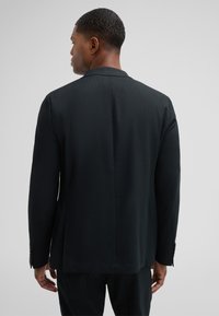 Schwarze maßgeschneiderte Anzugjacke, strukturierter Stoff, schlanker Schnitt, mit einem Schlitz hinten und geknöpften Ärmeln. Ansicht von hinten.