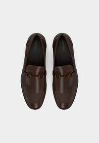 Mocassins en cuir brun à finition brillante, ornés d'une chaîne décorative de couleur or sur le dessus, avec un design à bout rond.