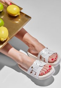 White woven sandals with thick platform soles, featuring a floral metal accent, paired with a hand holding a gold tray of lemons and limes.