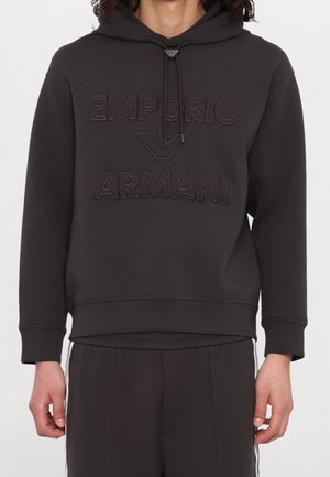 Musta huppari, jossa koholla oleva Emporio Armani -logo ja kiristysnauhallinen huppu, pukeutuneena henkilöllä, jolla on mustat housut valkoisilla sivuraitojen kanssa.