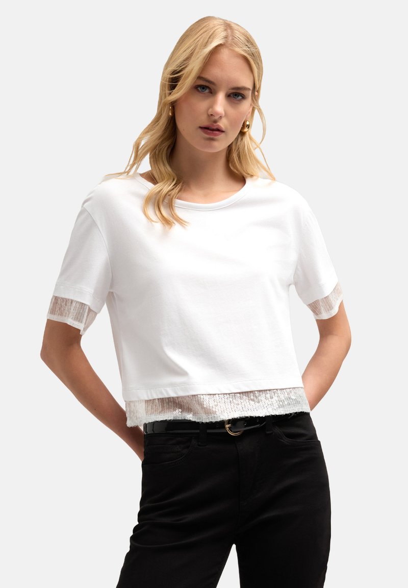 Hvid kortærmet crop top med gennemsigtig blondekant ved ærmerne og kanten, med rund halsudskæring og parret med sorte bukser.