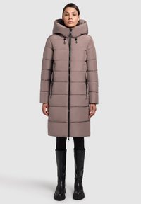 Puffercoat in lichtbruin met een capuchon; heeft een verticale gestikte patroon, een rits aan de voorkant en zijzakken. Gecombineerd met zwarte laarzen tot de knie.