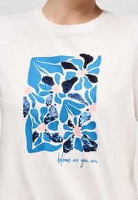 Λευκό μπλουζάκι με μπλε και ροζ αφηρημένο ανθικό σχέδιο που περιλαμβάνει μπλε παγιέτες και τη φράση «άνθισε όπως είσαι» με μπλε γραφικά γράμματα από κάτω.