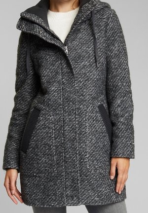 Femme portant un manteau en laine texturée gris foncé arrivant à mi-cuisse avec capuche, fermeture éclair et deux poches inclinées bordées de noir, par-dessus un pull blanc.