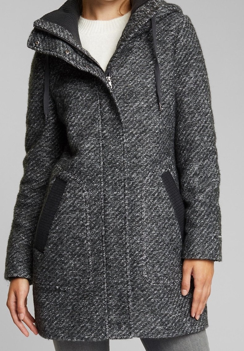 Femme portant un manteau en laine texturée gris foncé arrivant à mi-cuisse avec capuche, fermeture éclair et deux poches inclinées bordées de noir, par-dessus un pull blanc.