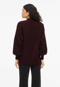 Vīna sarkanā adīta džempera augsts apkaklis, pūsts piedurknes un ribots aproču un apakšmalas dizains. Raksturīgs teksturēts materiāls un brīvs piegriezums.