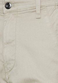 Pantalones de algodón beige con una textura suave, un cierre de botón negro y patrones de costura diagonal en los bolsillos delanteros.