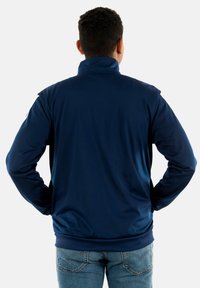 Navyblauwe jas met een hoge kraag, lange mouwen en een gladde textuur. Heeft witte accenten op de schouders en een aansluitende zoom.