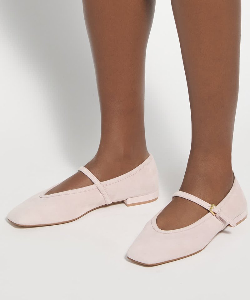 Chaussures plates Mary Jane rose clair avec bout carré et petite bride à boucle portées sur des jambes à la peau foncée.