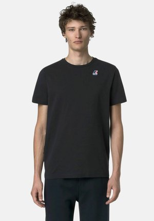 Giovane uomo che indossa una t-shirt nera semplice con un piccolo logo K-Way sul petto in alto a sinistra, in piedi su uno sfondo bianco.