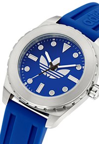 Ura z zifro modre silikonske naravnanice, ki vključuje ohišje iz nerjavečega jekla, živahno modro številčnico, izrazite bele oznake ur in logotip Adidas.