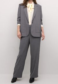 Costume gris sur mesure comprenant un blazer à un bouton avec deux poches à l'avant, associé à un pantalon à jambes larges et un chemisier crème avec un nœud.
