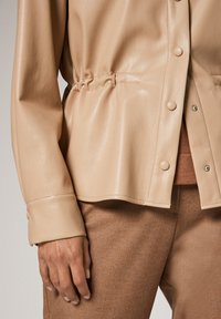 Veste en cuir beige courte avec des boutons-pression, détails de taille froncée et texture lisse, associée à un tissu texturé marron en dessous.