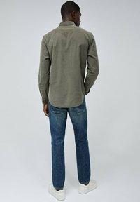 Camisa de manga larga verde olivo con dobladillo curvado, combinada con jeans de mezclilla azules y zapatillas blancas. La tela parece suave y casual.