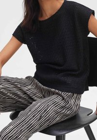 Top nero a maglia con maniche corte, caratterizzato da un motivo testurizzato. Abbinato a pantaloni a righe bianchi e neri, seduto su una sedia nera.