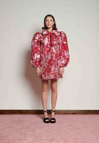 Ροζ λουλουδάτο φόρεμα με υπερμεγέθη μανίκια, μεγάλο φιόγκο στο γιακά και γυαλιστερό ύφασμα. Φορεμένο με μαύρες ψηλοτάκουνες πέδιλες με λουράκια σε φόντο ροζ μοκέτας.